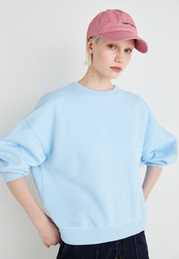 Sweatshirt oversized azul claro em material suave com punhos e bainha canelados, acompanhado de um boné rosa com um logótipo preto.