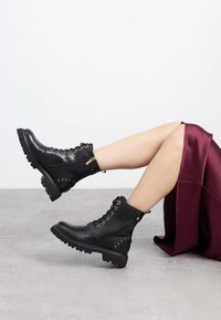 Botas de tobillo de cuero negro con diseño de cordones, ojales dorados, cremallera lateral y suela de goma gruesa con tachuelas en el lateral.