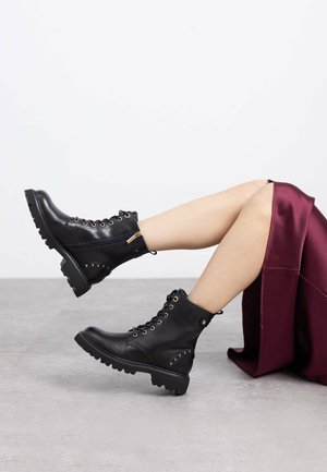 Botas de tobillo de cuero negro con diseño de cordones, ojales dorados, cremallera lateral y suela de goma gruesa con tachuelas en el lateral.