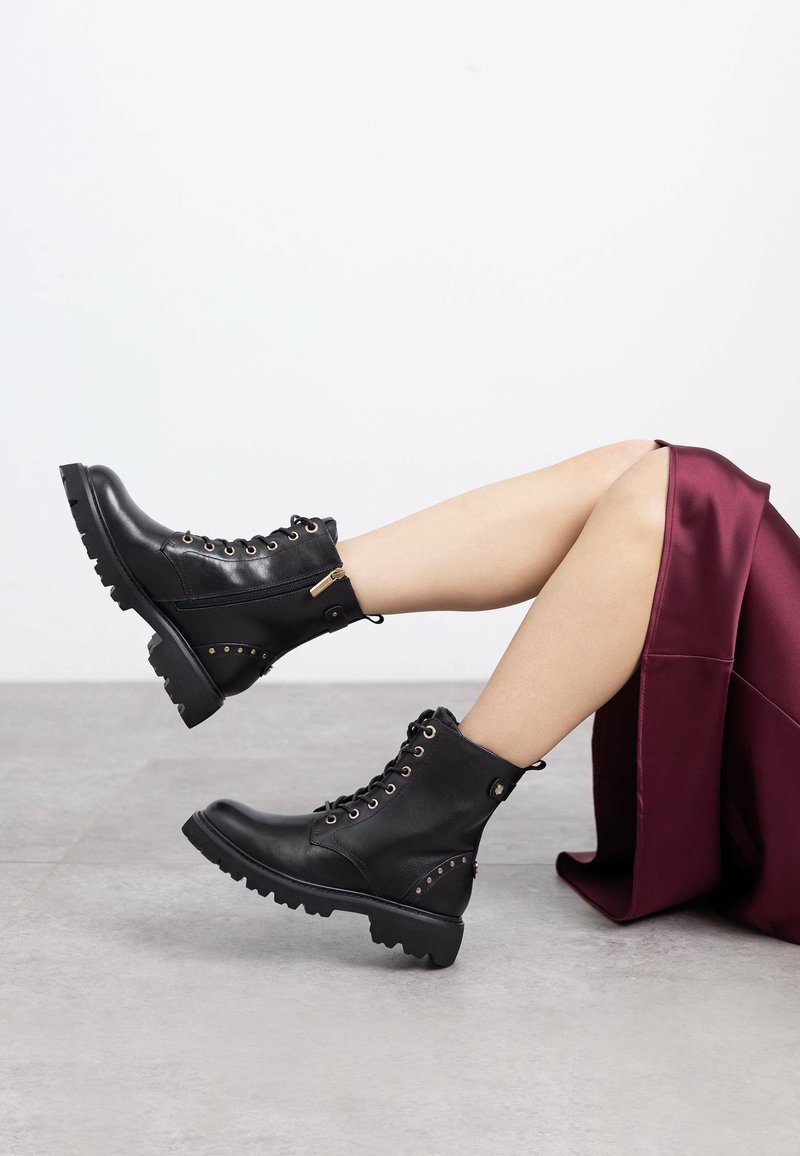 Botas de tobillo de cuero negro con diseño de cordones, ojales dorados, cremallera lateral y suela de goma gruesa con tachuelas en el lateral.