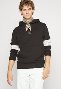 Schwarzer Hoodie mit weißen Ärmelakzenten und einer Fronttasche. Besitzt ein kleines Logo auf der Brust und einen beigen, gemusterten Schal um den Hals.