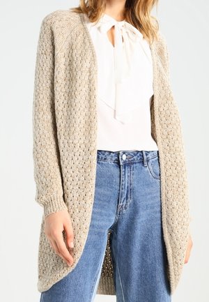 Kobieta ubrana w beżowy sweter ażurowy typu cardigan na białą bluzkę z kokardą, połączona z wysokim stanem niebieskich dżinsów.