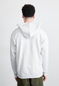Hombre con cabello rizado corto, vistiendo una sudadera gris claro y pantalones verde oscuro, de espaldas contra un fondo blanco liso.