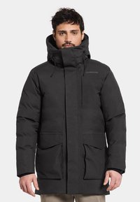 Parka nera isolata con colletto alto, cappuccio regolabile e due grandi tasche frontali. Presenta una superficie liscia e elementi di design minimali.