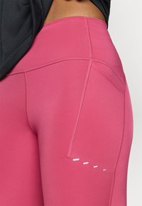 Siddende pink leggings med en glat tekstur, der har en sidelomme og kontrasterende hvide accentlinjer nær hoftområdet.