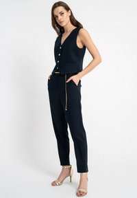 Granatowa bezrękawnik z złotymi guzikami, w połączeniu z tapered granatowymi spodniami. Złoty łańcuszek na spodniach. Modelka nosi złote szpilki.