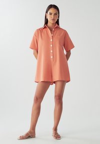 Korallfärgad kortärmad overall med krage, knappstängning framtill och en bröstficka. Mjukt tyg med avslappnad passform och shorts-liknande nederdel.