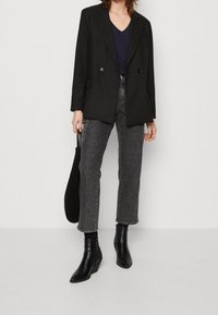 Schwarze zweireihige Blazerjacke mit Revers, getragen über einem navyfarbenen V-Ausschnitt-Oberteil, kombiniert mit hochgeschnittenen grauen Jeans und schwarzen Wedge-Ankle-Boots.
