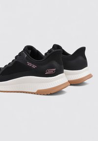 Baskets athlétiques noires avec texture en mesh, semelles intermédiaires blanches et semelles extérieures en caoutchouc. Caractérisées par le logo "Skechers Slip-Ins" en rose sur le côté.