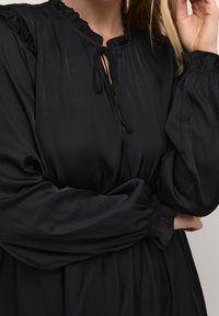 Svart flytande klänning med en volangkrage och långa, voluminösa ärmar. Tyget verkar slätt och något reflekterande med en knytning i midjan.