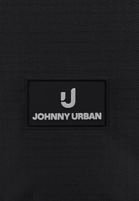 Sort stofbaggrund med en sort rektangulær label, der viser et hvidt "J" logo over teksten "JOHNNY URBAN" med hvide bogstaver.