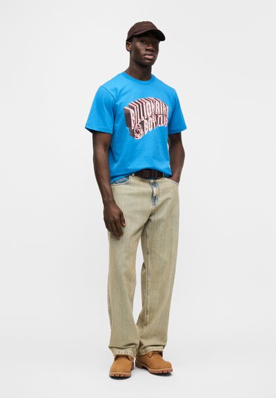 Μπλε βαμβακερό T-shirt με γραφικό σχέδιο, συνδυασμένο με ανοιχτόχρωμο τζιν παντελόνι με σχέδια και ταμπά καστόρινα μποτάκια. Το καφέ καπέλο ολοκληρώνει την εμφάνιση.