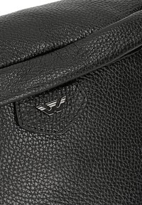 Borsa in pelle nera con superficie testurizzata, chiusura a patta e un emblema di marca metallico con contorno esagonale.