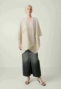 Top estilo quimono bege com detalhes em franjas ao centro, sobreposto a uma saia maxi de ganga escura, combinado com sandálias pretas.