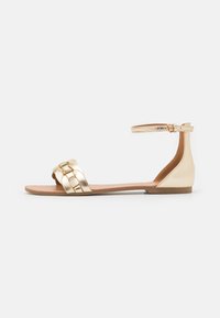 Gold metallic flat sandal with a braided strap design, ankle buckle closure, and a smooth, brown sole.