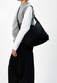 Černá kabelka přes rameno z texturovaného materiálu; má zakřivený tvar, nastavitelný popruh a zapínání na zip. Nositelná pod šedou vestou.