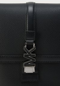 Μαύρη δερμάτινη τσάντα με υφή, που διαθέτει κομψό λουράκι και μεταλλικό κουμπί με λογότυπο και γεωμετρικά διακοσμητικά στοιχεία.