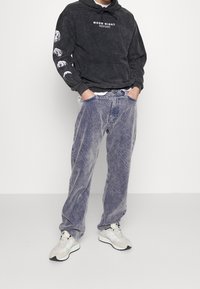 Uomo che indossa una felpa nera con fasi lunari sulla manica e scritta "Moon Night", pantaloni di velluto a coste blu sbiadito e scarpe da ginnastica beige, con le mani nelle tasche.