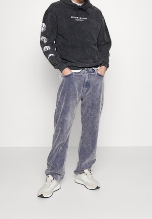 Homme portant un sweat à capuche noir avec des phases de lune sur la manche et le texte « Moon Night », un pantalon en velours côtelé bleu délavé et des baskets beige, les mains dans les poches.