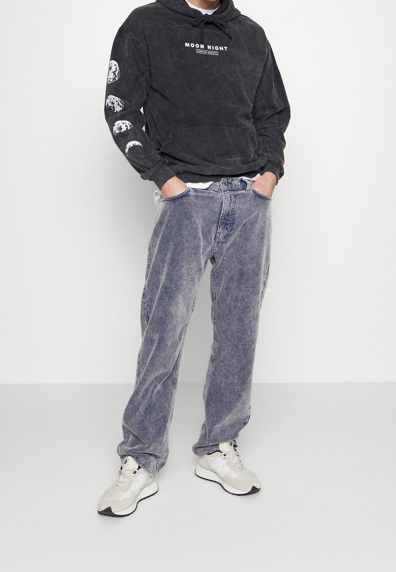 Uomo che indossa una felpa nera con fasi lunari sulla manica e scritta "Moon Night", pantaloni di velluto a coste blu sbiadito e scarpe da ginnastica beige, con le mani nelle tasche.