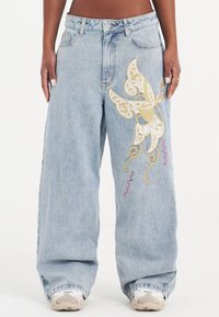 Lichtblauwe wijduitbroek heeft gouden en roze vlinderborduring met spiralen, gemaakt van denim met een zachte, versleten textuur.