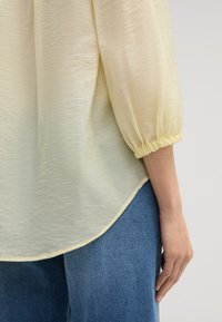 Blusa gialla chiaro con tessuto strutturato, maniche a palloncino raccolte ai polsi e orlo curvo, abbinata a jeans blu.