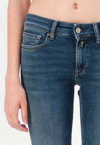 Tuvumā redzams cilvēks, kas valkā zila džinsa bikses ar pogu aizdari, rādot vidukli, kabatas un daļu no kreisās rokas.