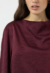 Burgundská texturovaná blůza s širokým výstřihem, vystavující zmačkanou látku. Materiál vypadá hladce s jemnými variacemi v vzoru.