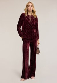 Sukienka z aksamitu w kolorze burgundowym, z podwójnym zapięciem i przednimi kieszeniami, oraz szerokimi spodniami. Towarzyszy jej mała brązowa torebka.