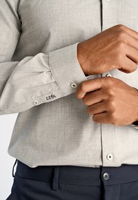 Hellgraues, strukturiertes Button-up-Hemd mit spitzem Kragen, Knöpfen an den Manschetten und genähten Details an den Ärmeln. Tragen mit dunklen Hosen.