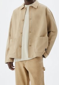 Lys beige jakke lavet af bomuldsblanding med knaplukning foran, to store lommer og en afslappet pasform, kombineret med en lysefarvet skjorte under.