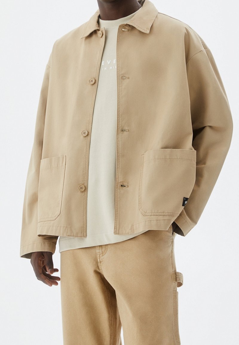 Lys beige jakke lavet af bomuldsblanding med knaplukning foran, to store lommer og en afslappet pasform, kombineret med en lysefarvet skjorte under.