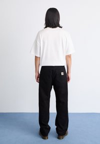 Camiseta blanca de gran tamaño con mangas cortas combinada con pantalones negros que presentan una etiqueta en el bolsillo trasero. Ambientada sobre un fondo claro.