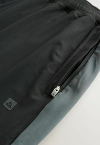 Black athletic shorts with a gray waistband, featuring a side zip pocket and a textured fabric. Accented with a logo patch near the hem.