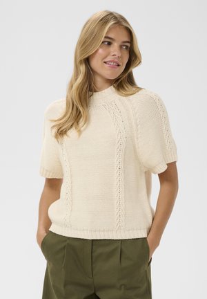 Frau mit langen blonden Haaren, die einen cremefarbenen kurzärmligen Strickpullover und olivgrüne Hosen trägt, Hände in den Taschen, blickt zur Seite und lächelt.
