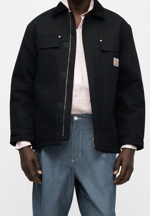 Mann trägt schwarze Jacke mit Reißverschluss und zwei Brusttaschen über einem hellrosa Hemd mit Knöpfen und lockeren blauen Jeans.