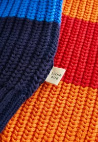 Coperta lavorata a maglia con strisce blu, blu navy, rosse e arancioni. Include un piccolo cartellino etichettato 'Little Bird'. Materiale in filato spesso e testurizzato.