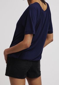 Mörkblå kortärmad topp med avslappnad passform, med rund halsringning och rynkade detaljer på baksidan, tillsammans med svarta denimshorts.