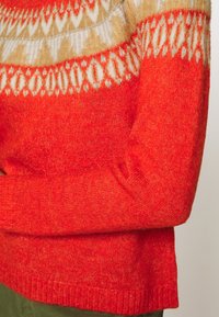 Röd tröja med en texturerad stickning och en gradient från orange till kräm vid halsringningen, accentuerad med silvertråd.