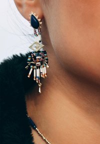 Boucles d'oreilles en perles avec un accent doré, présentant une pierre en forme de goutte d'eau bleue, une forme de diamant, et des franges de couleurs variées.