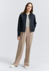 Navy zip jacket with a collar, paired with a striped top, beige wide-leg trousers, and white sneakers. Smooth fabric and tailored fit.