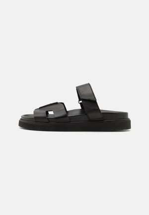 Black leather slide sandals with flat soles, wide straps, and a square cutout detail. Textured and smooth surface finishes.