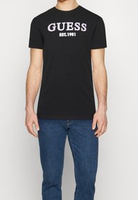 Sort bomuld t-shirt med "GUESS" i fed, pink skrifttype og "EST. 1981" nedenunder. Rund halsudskæring med korte ærmer.