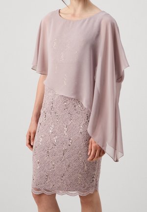 Frau trägt ein hell mauvefarbenes Kleid mit einem Spitzen-Paillettenrock und einem transparenten asymmetrischen Cape-Oberteil mit weiten Ärmeln.