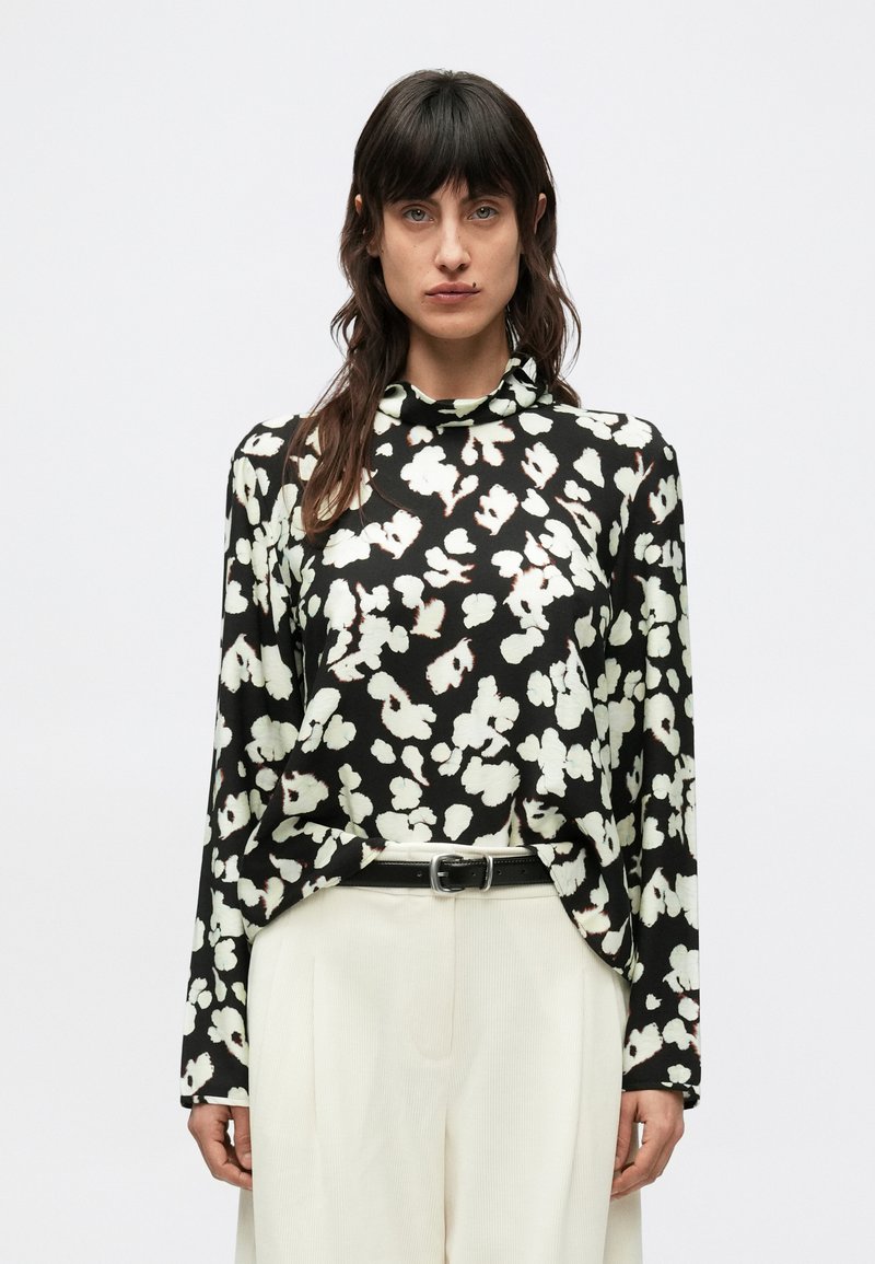 Czarna bluzka z długim rękawem w kremowy kwiatowy wzór, z wysokim kołnierzem i luźnym krojem, zestawiona z jasnymi, plisowanymi spodniami.