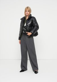 Černá péřová bunda s lesklým povrchem, široké šedé kalhoty a černá taška. Modelka stojí na bílém pozadí a předvádí outfit.