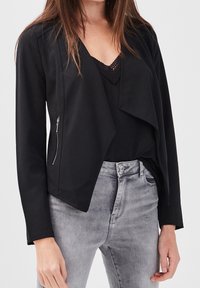 Veste légère noire avec un devant ouvert, un drapé subtil, des manches longues et un détail de poche zippée du côté gauche.