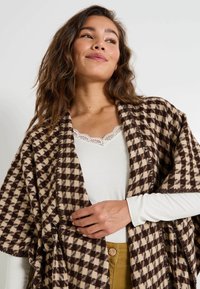 Châle à carreaux brun et crème avec une texture douce, porté sur un haut blanc. Présente un devant ouvert et un design décontracté.