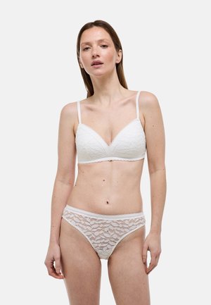 Femme aux cheveux raides portant un soutien-gorge en dentelle blanche et une culotte assortie en dentelle, debout devant un fond clair uni.