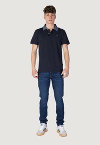 Polo navy con colletto azzurro, pattina con tre bottoni, indossato con jeans blu e sneakers bianche con dettagli verdi.